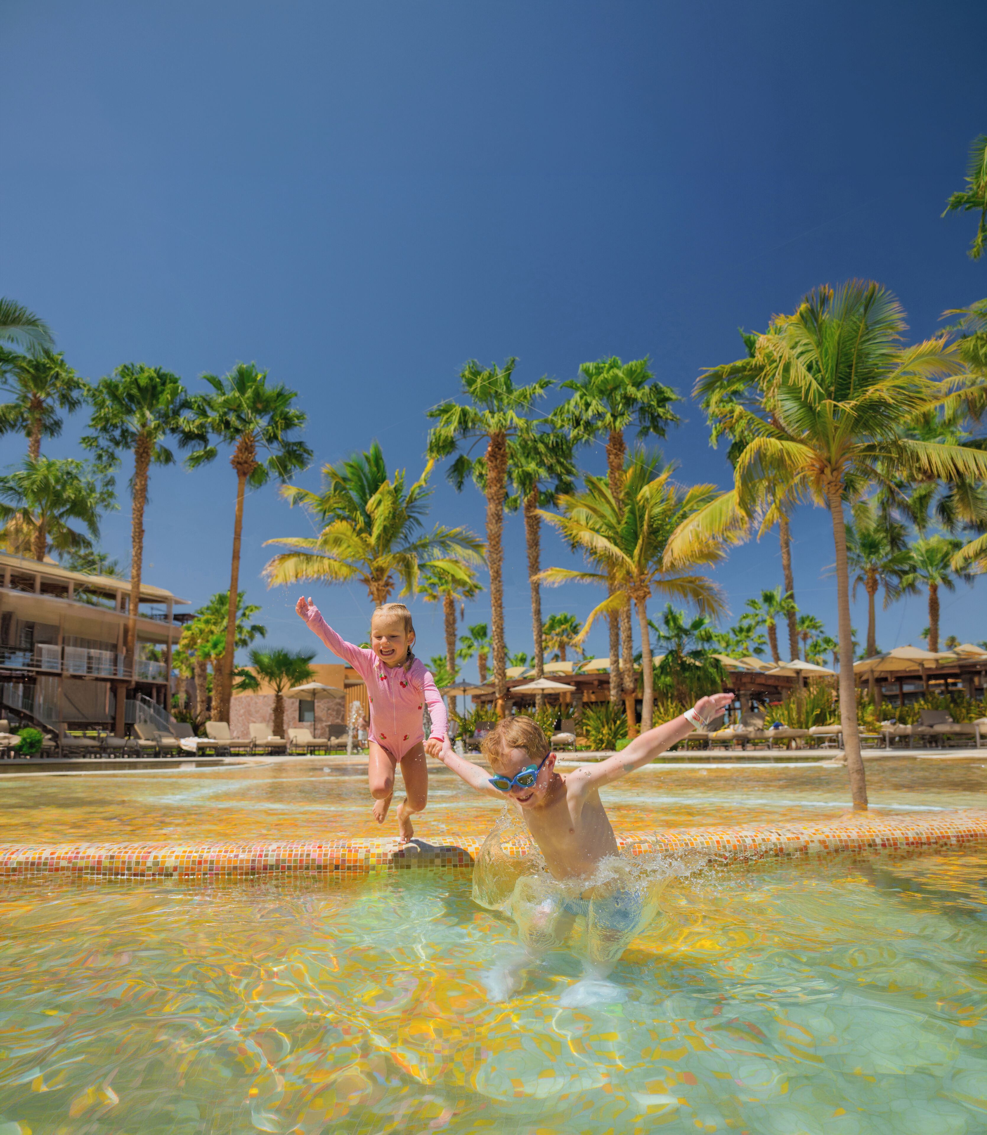 A swimming pool with clear water, surrounded by palm trees and a resort building.
