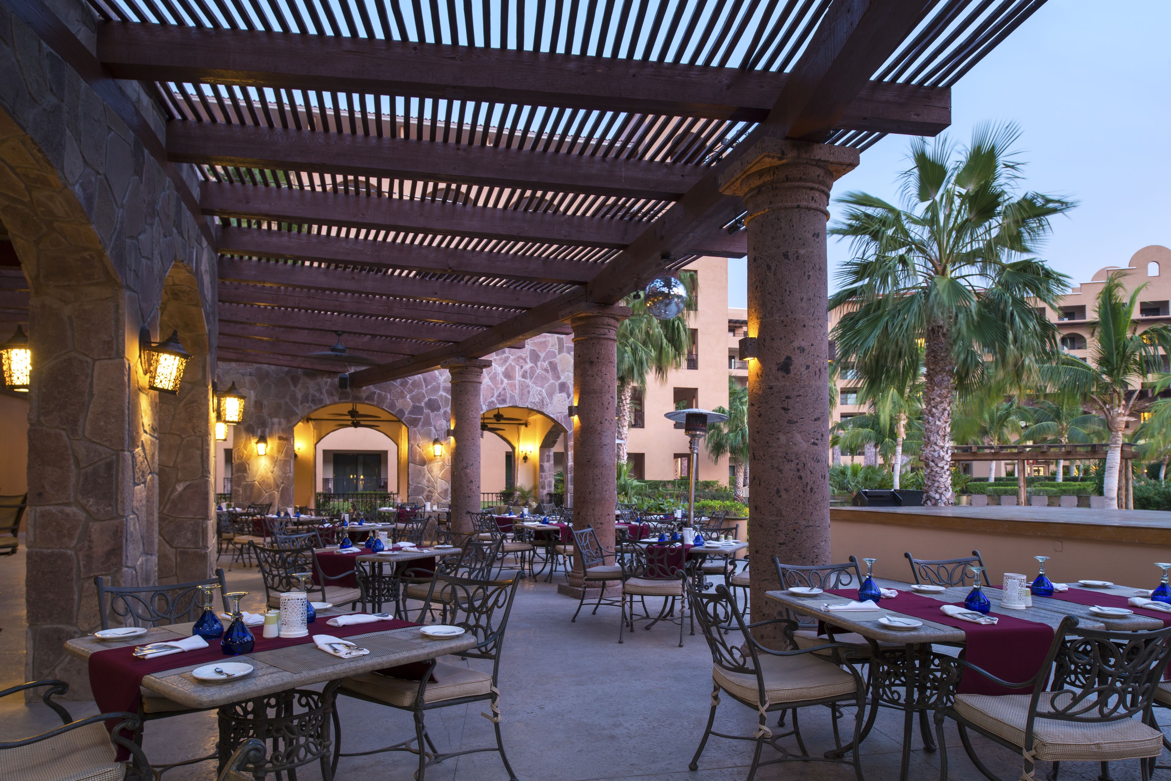 An outdoor dining area with stone pillars, wooden pergola, and tables set for dining.