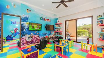 Une salle de jeux pour enfants avec un sol coloré, un ventilateur de plafond, une télévision, ainsi que divers jouets et jeux.