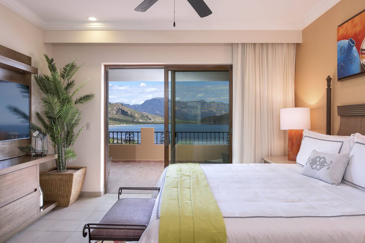 Ein Hotelzimmer mit Bett, Fernseher, Pflanze und einem Balkon mit Blick auf Berge und Wasser.