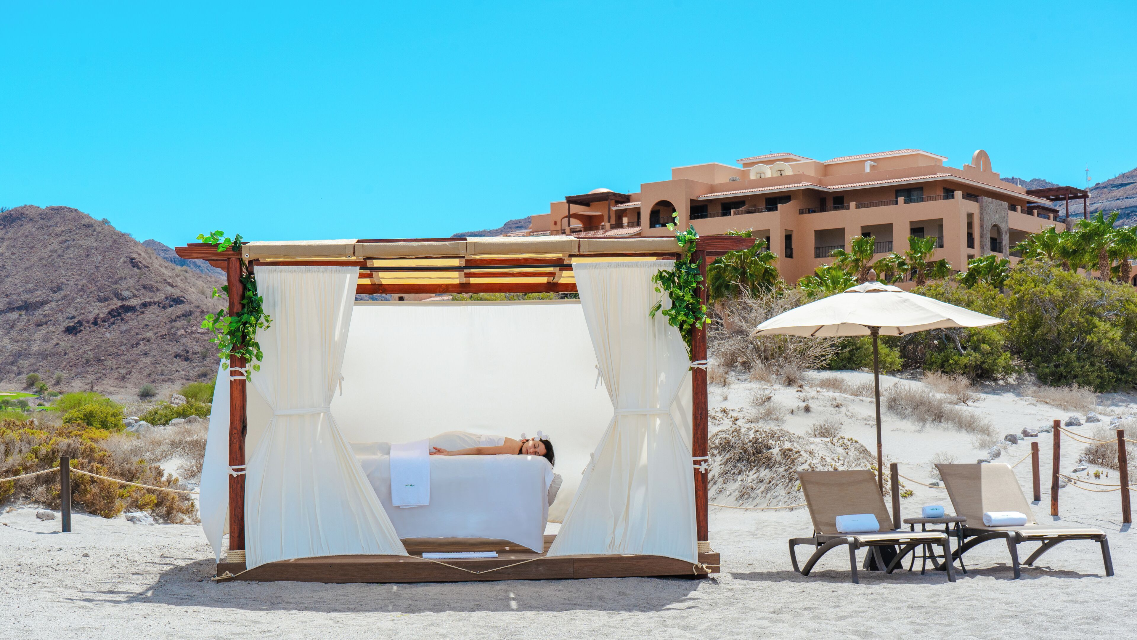 A beach cabana with a bed, two chairs, and an umbrella. It is located on a sandy beach with a mountain in the background and a building with balconies.