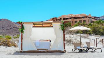 A beach cabana with a bed, two chairs, and an umbrella. It is located on a sandy beach with a mountain in the background and a building with balconies.