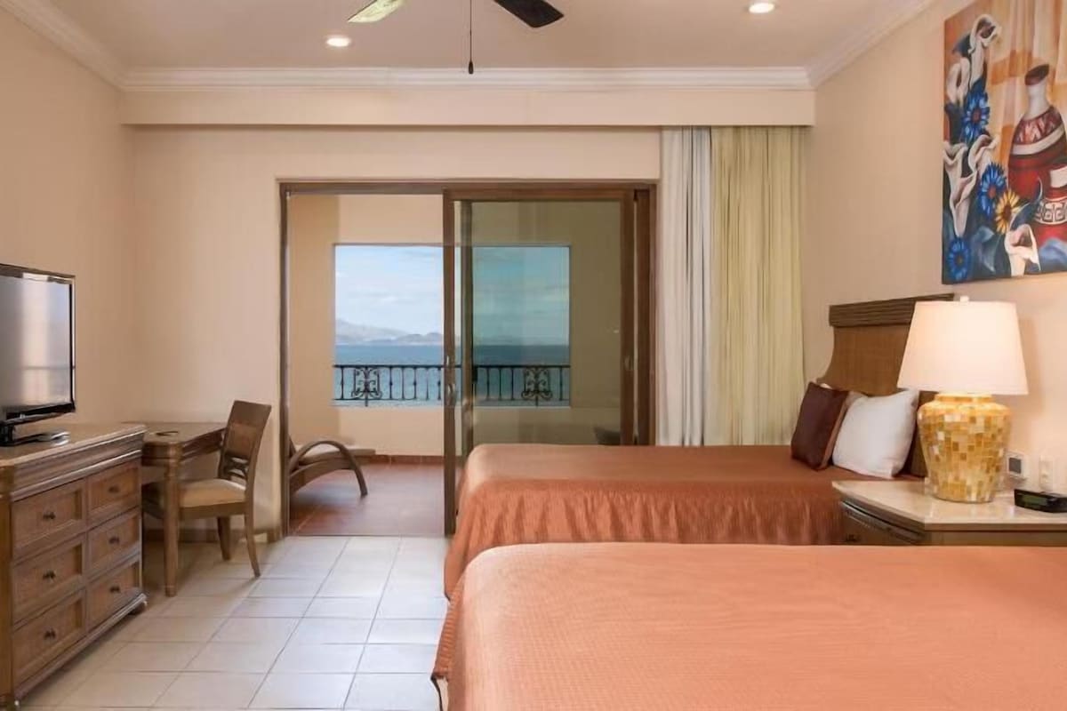 Hotelkamer met een bed, een bureau met een televisie, een stoel, een balkon met uitzicht en een schilderij aan de muur.