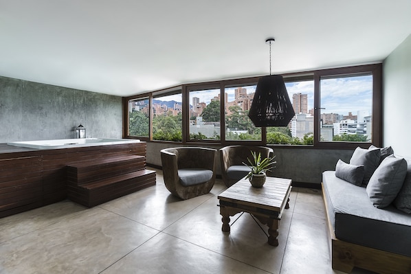A modern living room with a large window, a built-in hot tub, two armchairs, a sofa, and a wooden coffee table. - The Charlee Hotel (Medellín)