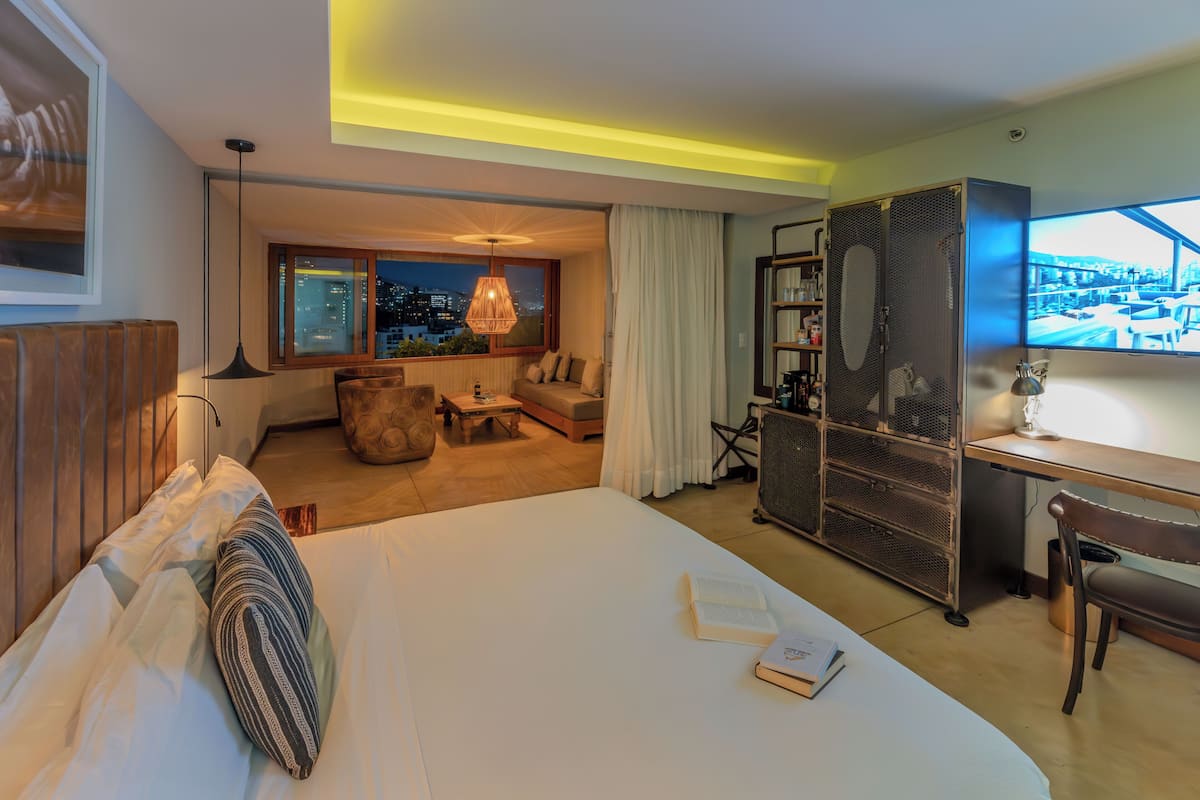 Ein Hotelzimmer mit einem großen Bett, einem Schreibtisch, einem Sessel, einem Bücherregal und Blick auf die beleuchtete Stadt.