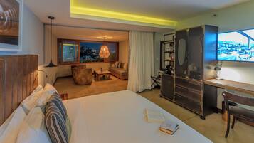 Habitación de hotel con una cama grande, un escritorio, una silla, una estantería y vistas de la ciudad de noche.