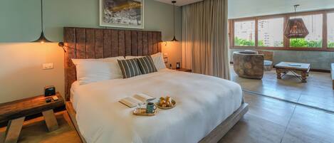 A spacious bedroom with a large bed, bedside tables, a reading area with a chair and table, and a view of greenery through large windows.