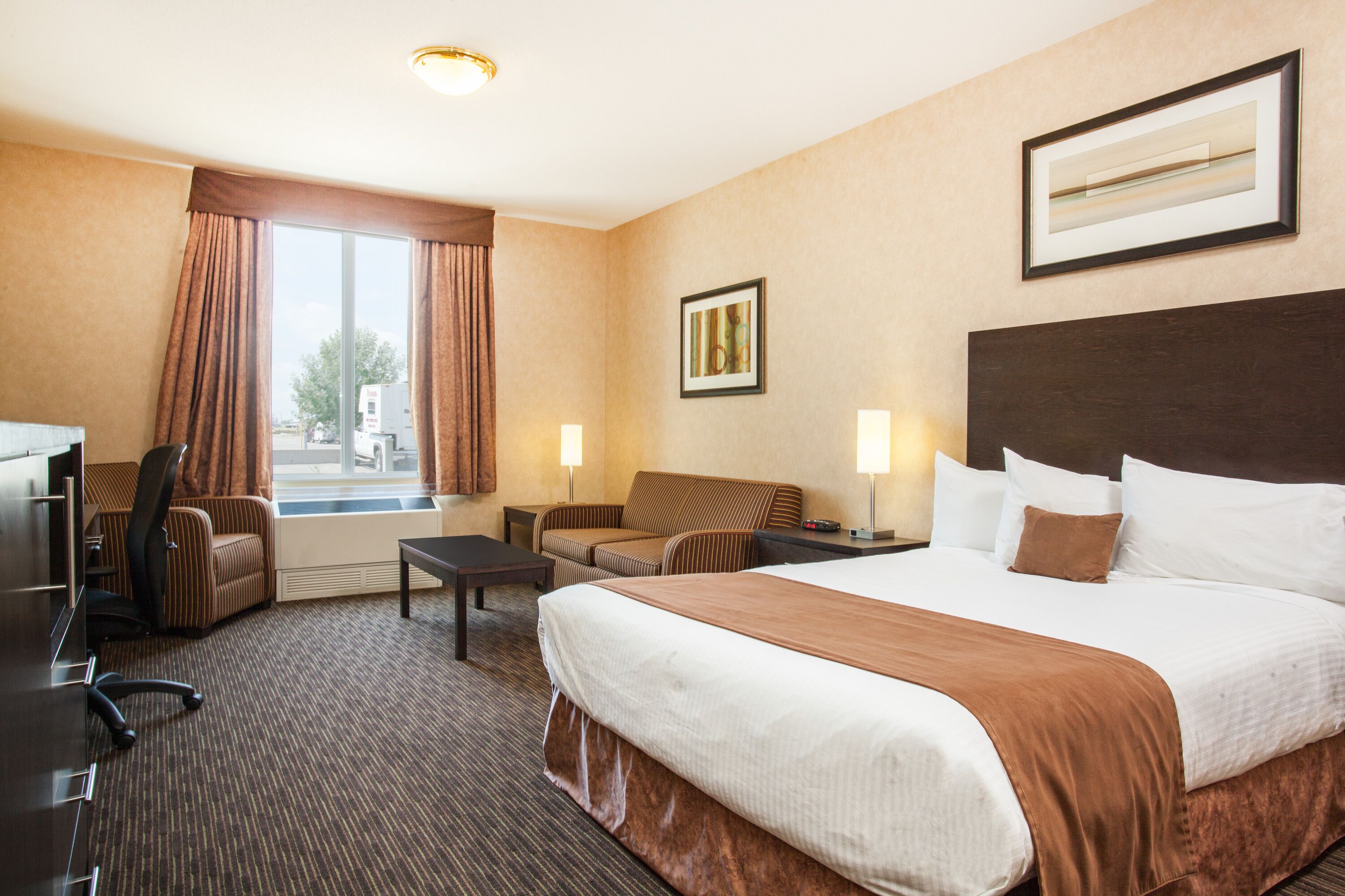 Habitación de hotel con una cama grande, un escritorio, una silla y un sofá. Se ve una ventana con cortinas, un cuadro en la pared y una lámpara en el escritorio.