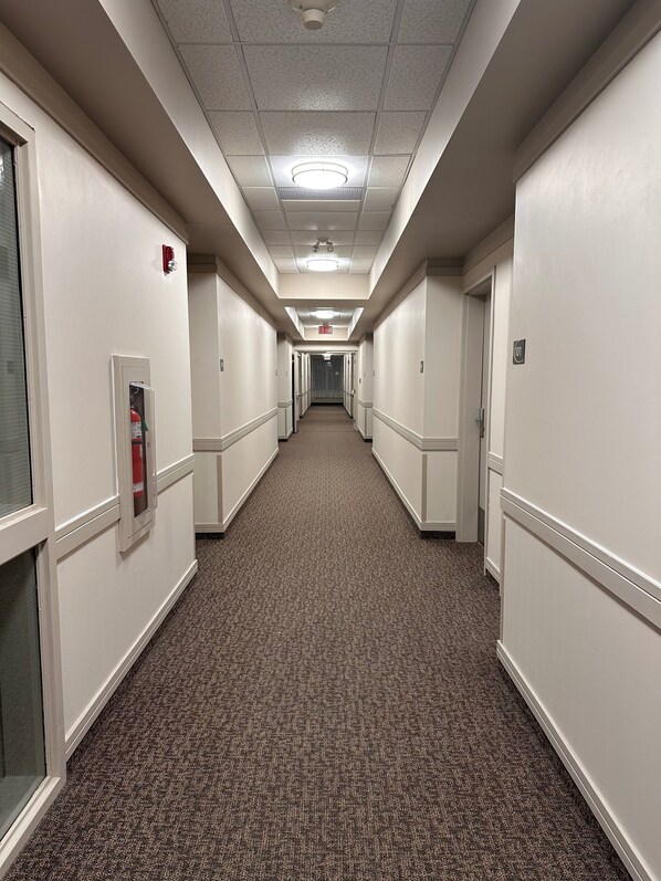 A hotel hallway with carpeted floors, white walls, and doors on both sides. - Ramada by Wyndham Estevan (Estevan)