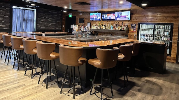 A bar area with a long counter, multiple stools, a television mounted on the wall, and a refrigerator stocked with beverages. - Ramada by Wyndham Estevan (Estevan)
