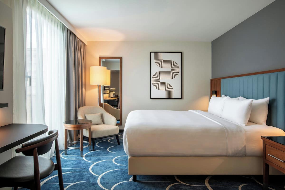 Quarto de hotel com cama, escrivaninha, cadeira, abajur e uma obra de arte na parede.