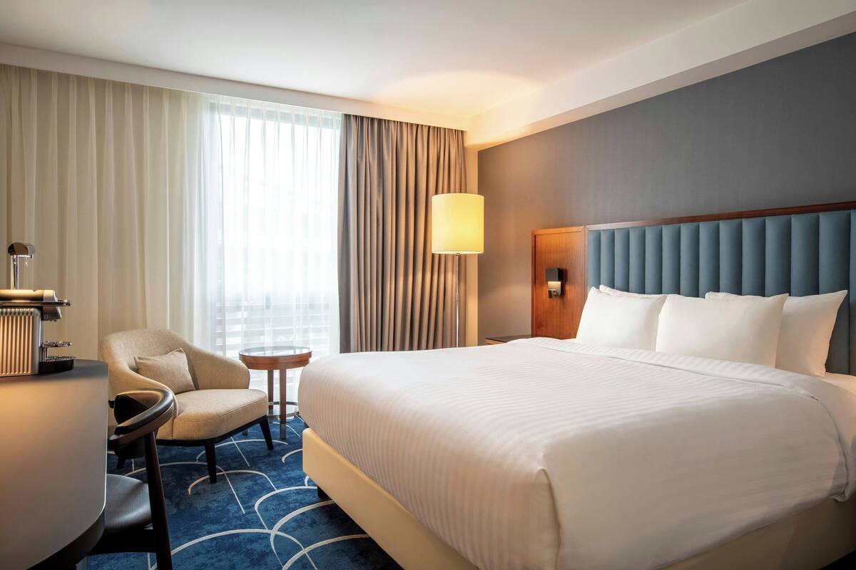 Quarto de hotel com uma cama grande, uma escrivaninha, uma cadeira, um abajur e vista para o exterior pela janela.