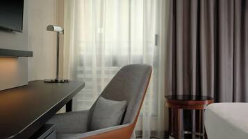 Ein Hotelzimmer mit einem modernen Sessel, einem kleinen runden Tisch, einem an der Wand montierten Fernseher und einem großen Fenster mit Vorhängen.