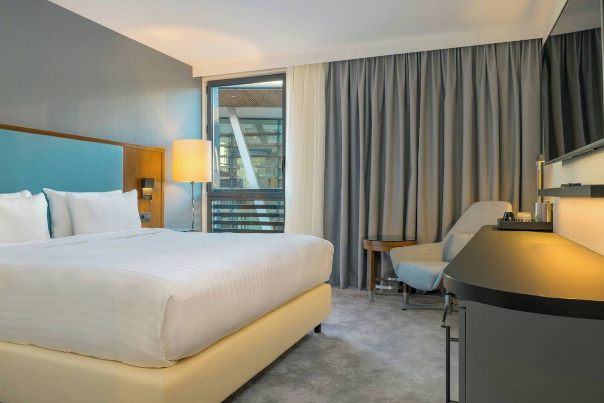 Quarto de hotel com uma cama grande, uma escrivaninha, uma cadeira, um abajur e vista para um edifício através de uma janela.