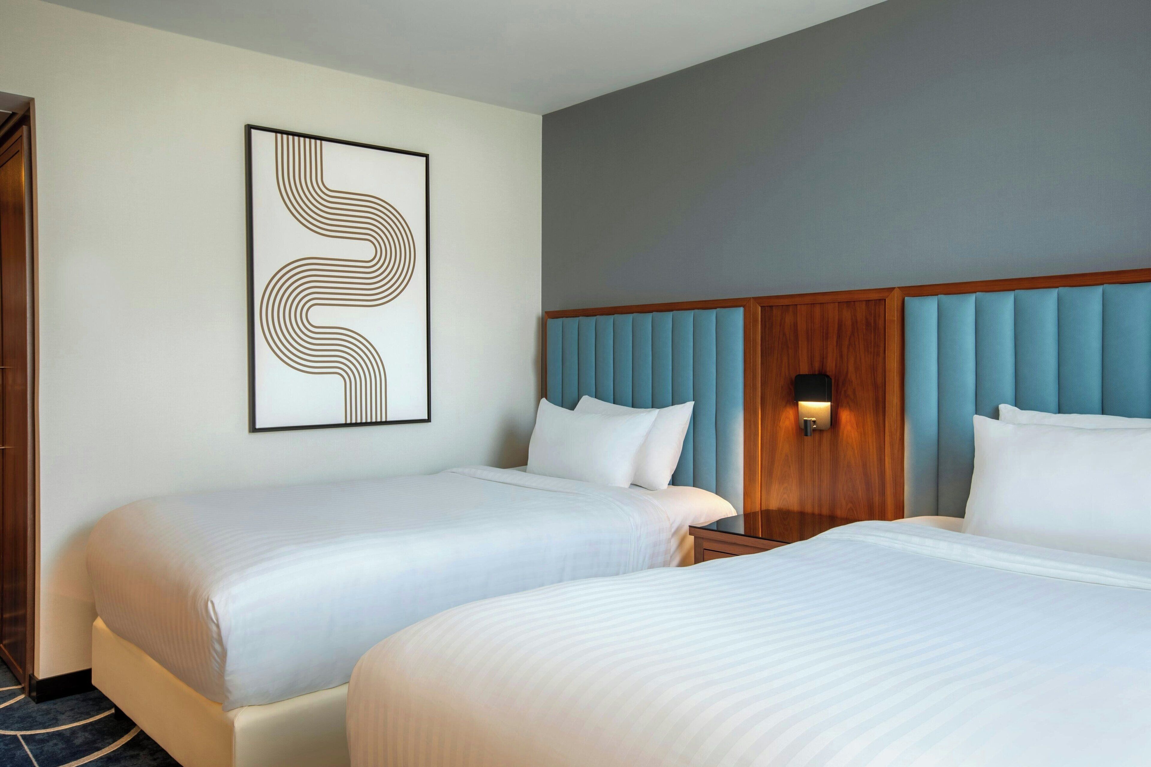 Ein Hotelzimmer mit zwei Betten, einem Holzkopfende, einem an der Wand befestigten Kunstwerk und einem Nachttisch mit Lampe.