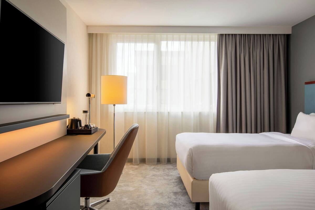 Quarto de hotel com cama, escrivaninha, cadeira, televisão e luminária.