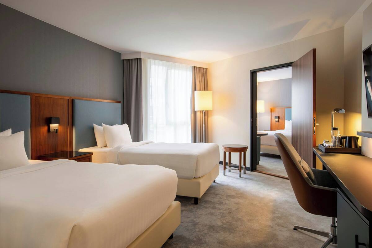 Quarto de hotel com duas camas, uma escrivaninha, uma cadeira, uma luminária e vista para o corredor.