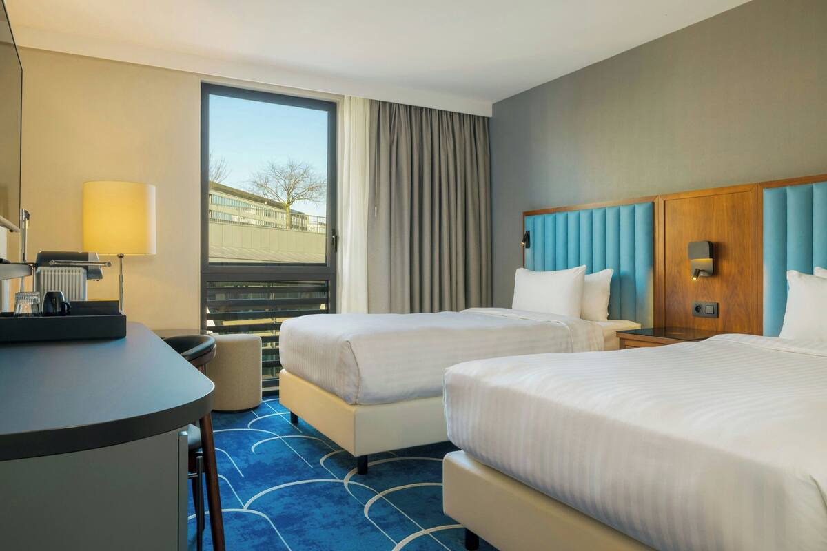 Quarto de hotel com duas camas, uma escrivaninha, uma cadeira, um abajur e uma janela com cortinas.