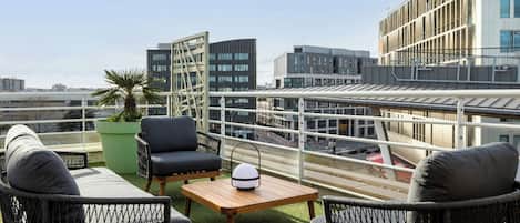 A rooftop terrace with wicker furniture, a green potted plant, and a view of modern buildings.