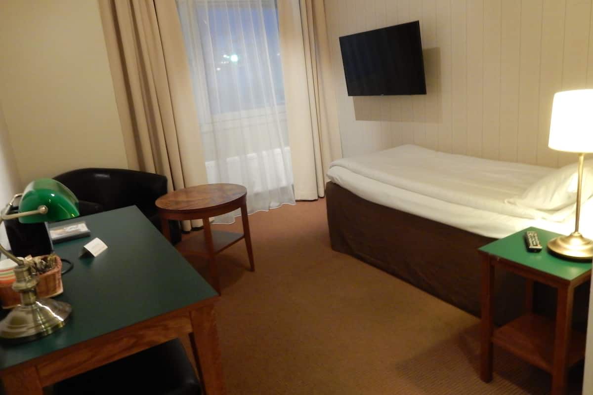Une chambre d’hôtel avec un lit, un bureau avec une lampe, une chaise, un téléviseur fixé au mur et une fenêtre avec des rideaux.