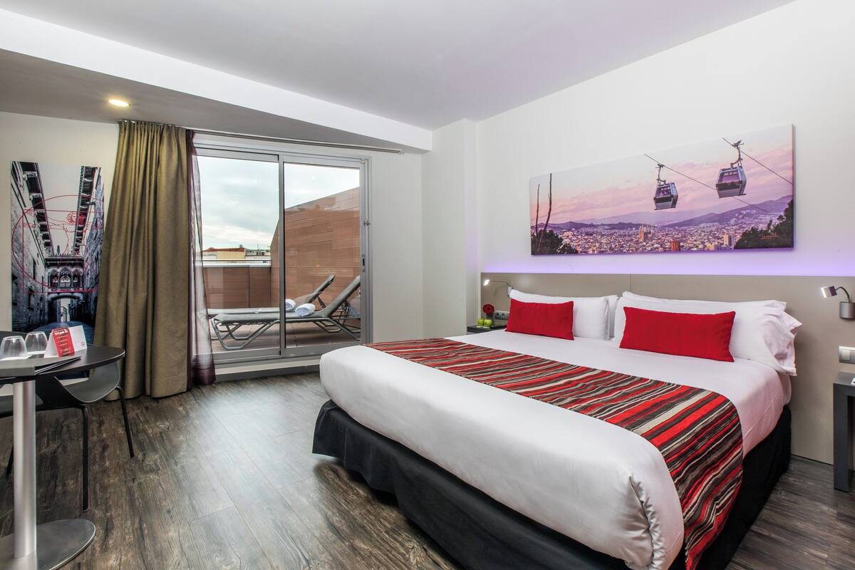 A hotel room with a large bed, a desk with a chair, a balcony with a view of the city, and a painting of a cityscape with cable cars.