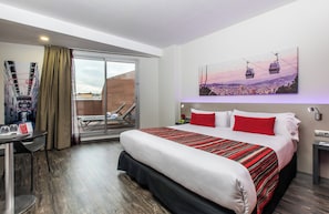 A hotel room with a large bed, a desk with a chair, a balcony with a view of the city, and a painting of a cityscape with cable cars. - Leonardo Boutique Hotel Barcelona Sagrada Familia (Barcelona)