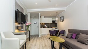 A modern living room with a sofa, a coffee table, and a TV mounted on the wall. There is a kitchen area with cabinets and a microwave.