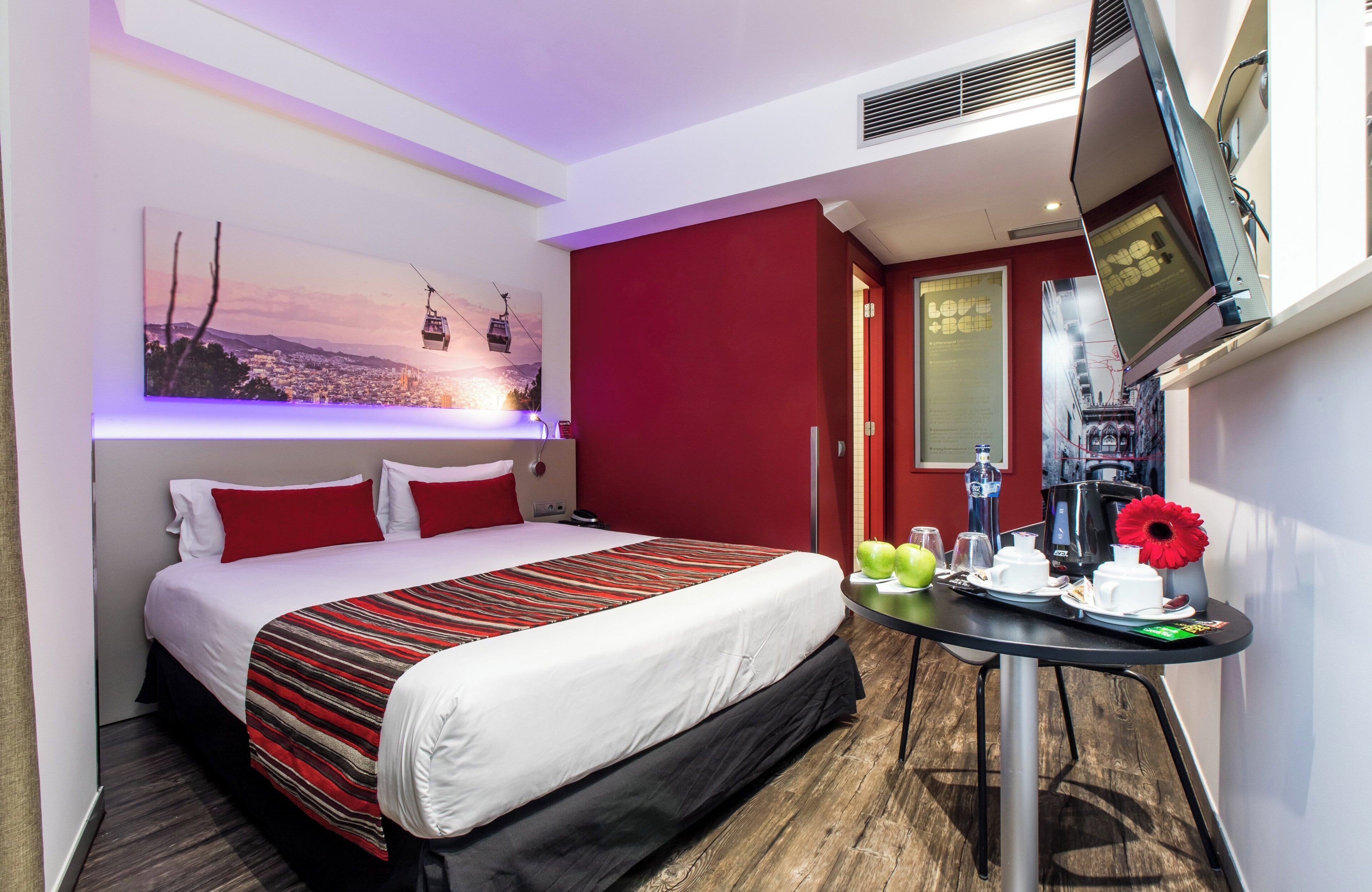 A hotel room with a bed, a TV mounted on the wall, a table with breakfast items, and a painting of a cable car on the wall.