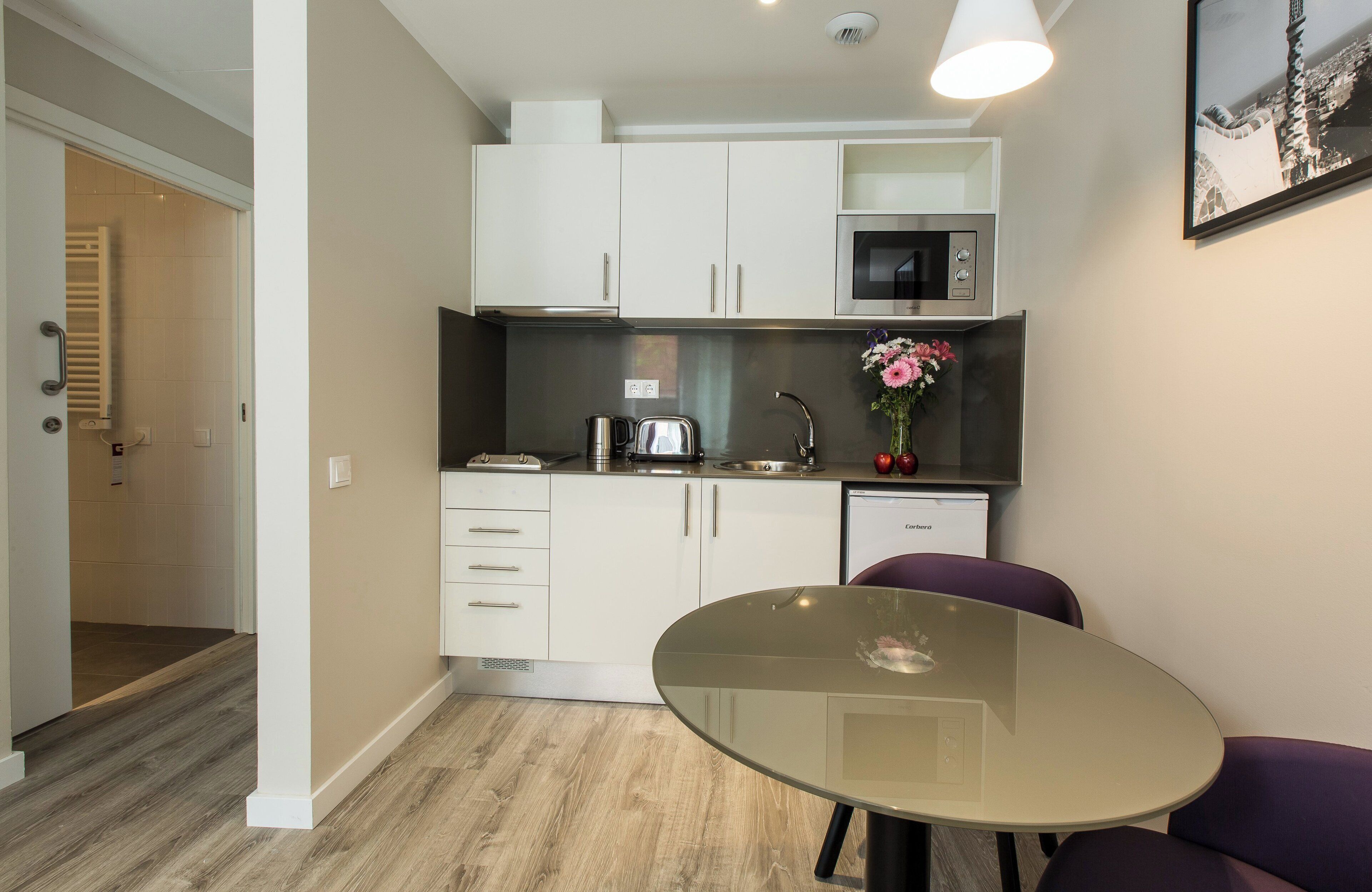 Una cocina compacta con armarios blancos, microondas, cafetera y una pequeña mesa de comedor con sillas moradas.