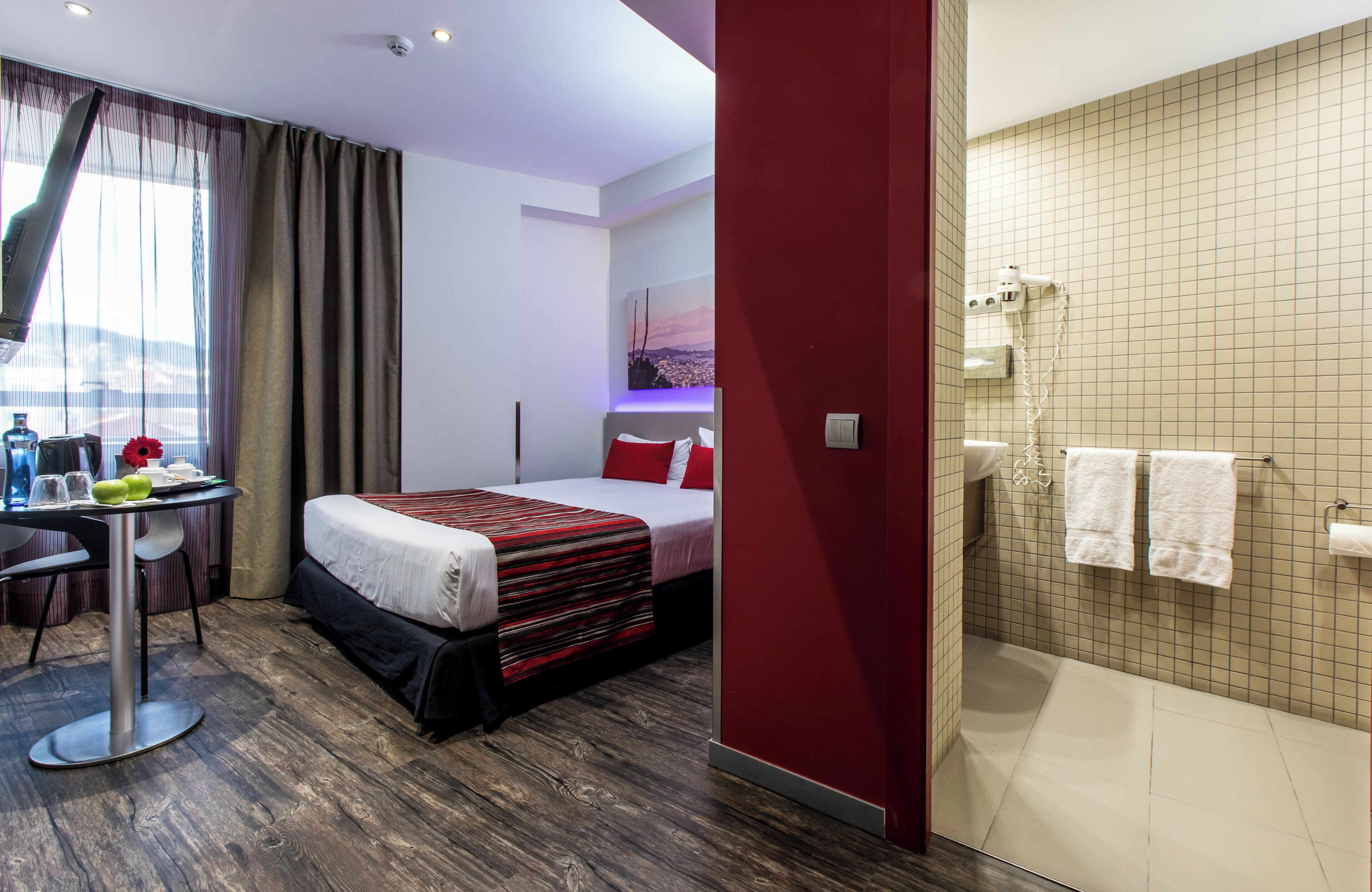 A hotel room with a bed, a desk with a chair, a shower area, and a window with curtains.
