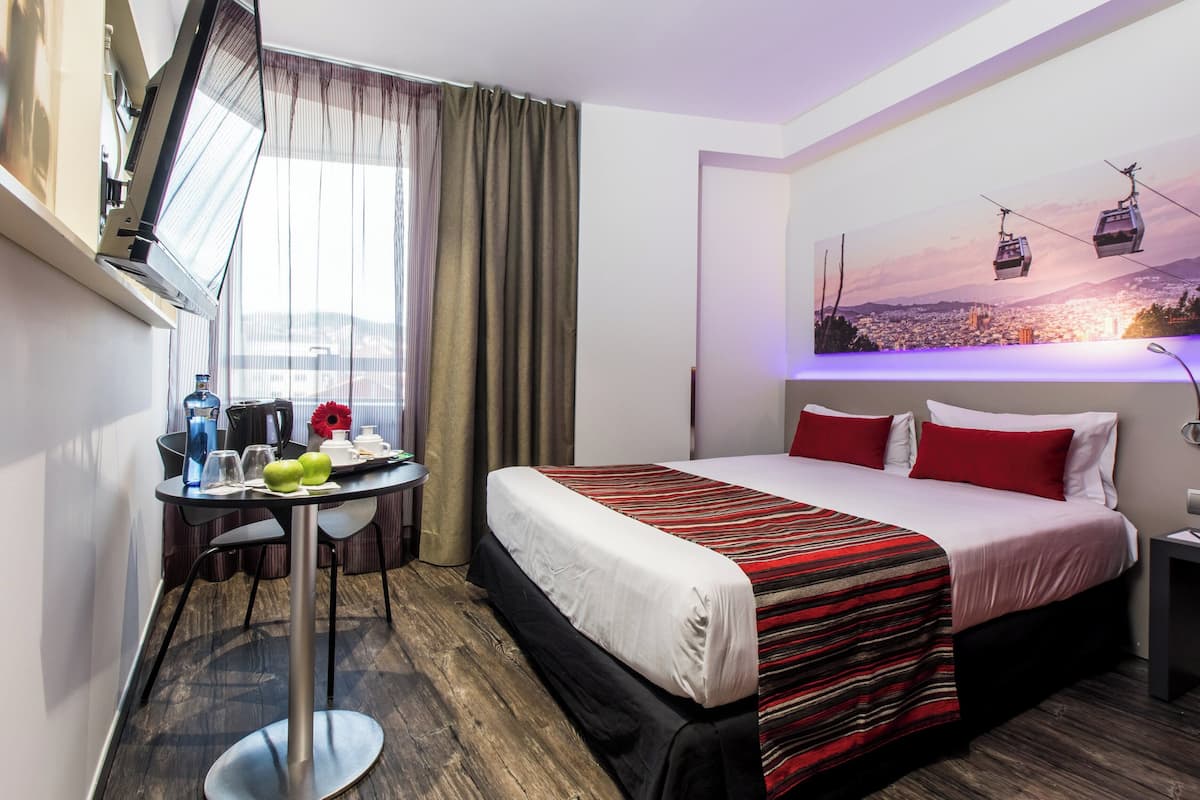 Ein Hotelzimmer mit einem Bett, einem Schreibtisch mit Stuhl, einem Fenster mit Vorhängen und einem an der Wand befestigten Fernseher.