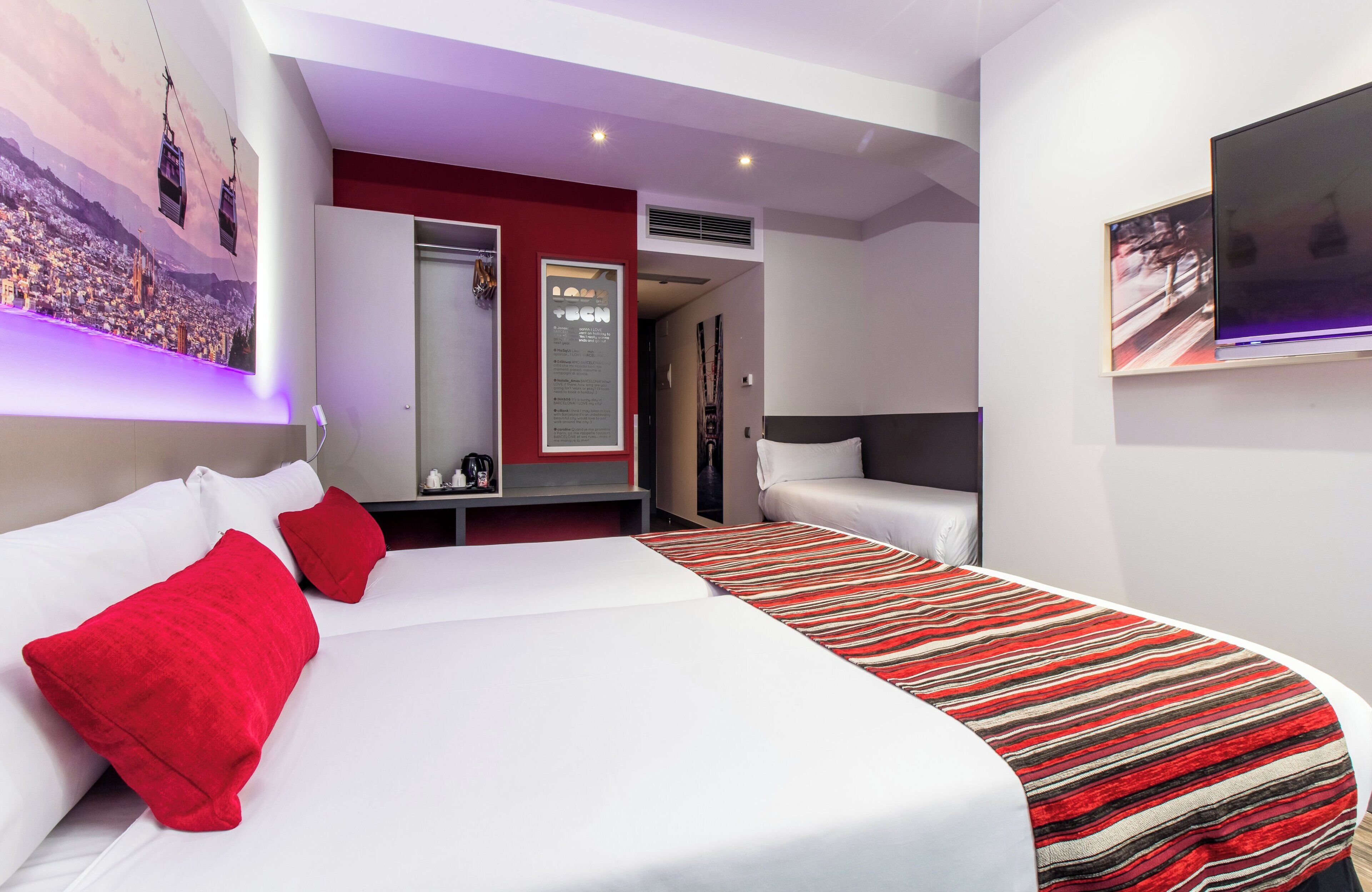 Una habitación de hotel moderna con una cama grande, almohadas rojas, una manta de rayas y un televisor montado, además de un mural con vista a la ciudad.