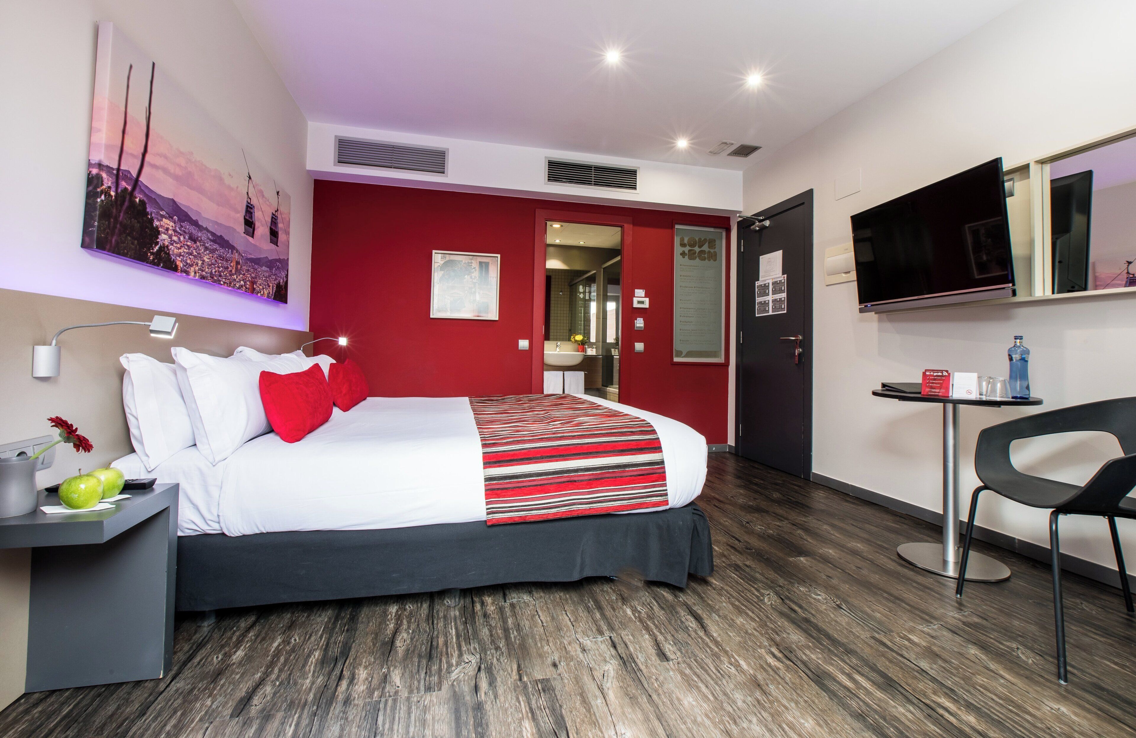 Habitación de hotel moderna con una cama grande, un escritorio con lámpara, televisor y una pared decorada en rojo.