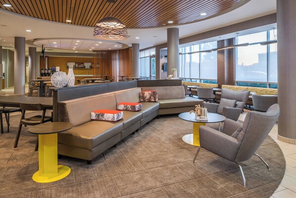 A modern lounge area with a curved wooden ceiling, a seating area with sofas and armchairs, a round table with chairs, and a large window providing a view of the city. - SpringHill Suites Huntsville Downtown (Huntsville)