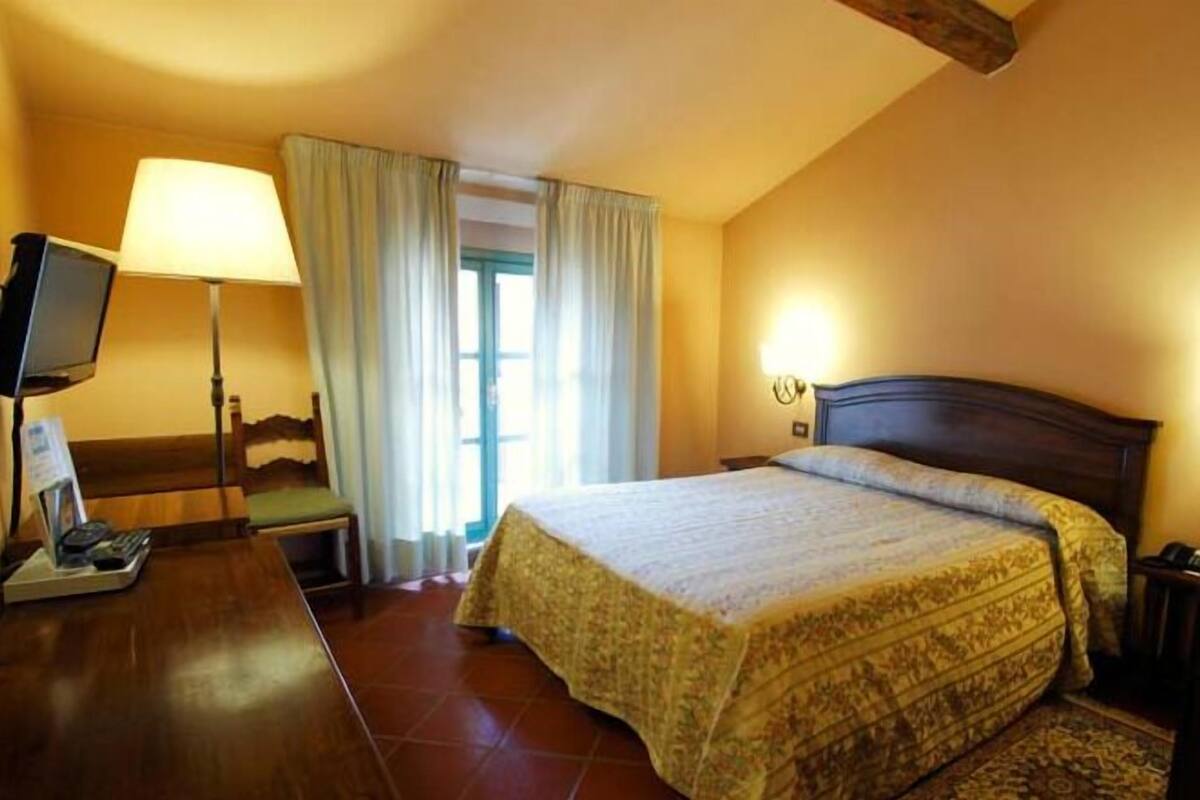 Une chambre d’hôtel avec un lit, un bureau avec une lampe, une télévision et une fenêtre avec des rideaux.