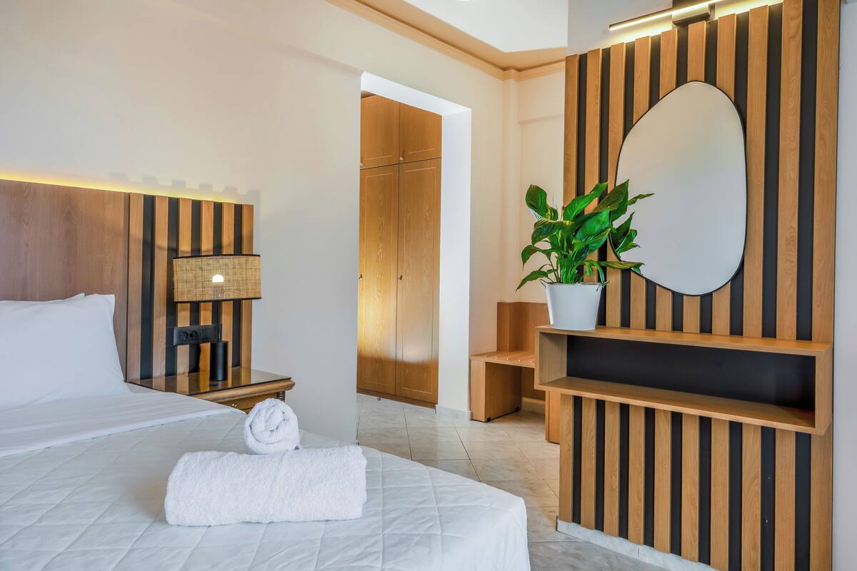 A hotel room with a large bed, a bedside table with a lamp, a mirror, a plant, and a wooden wardrobe.