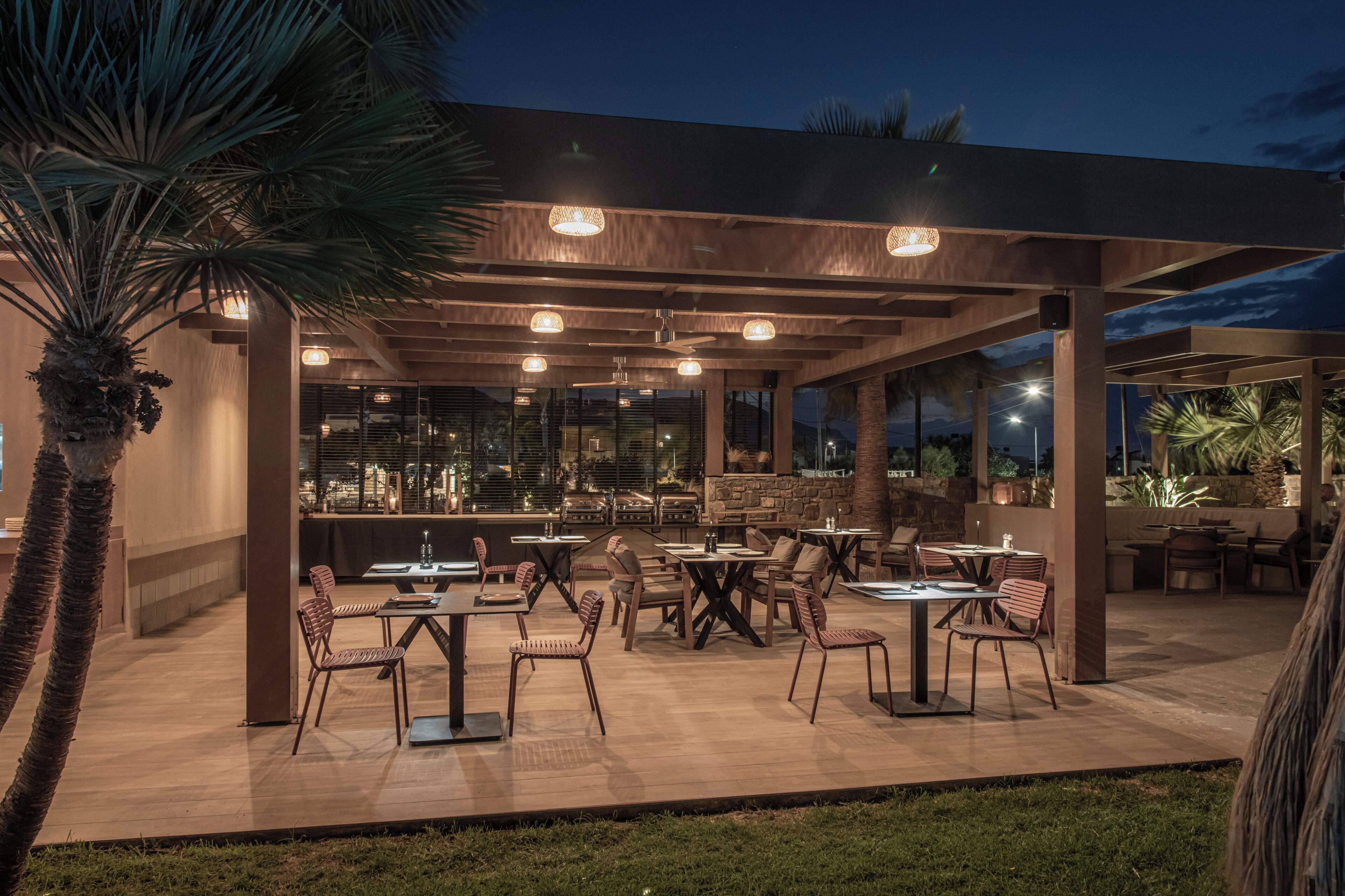 An outdoor patio area with tables and chairs, palm trees, and a covered seating area with hanging lights.