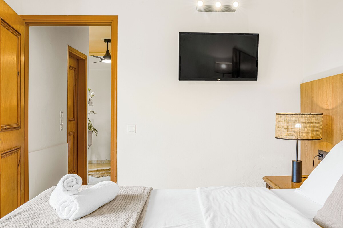 A hotel room with a bed, a TV mounted on the wall, a lamp on a bedside table, and a wooden door.