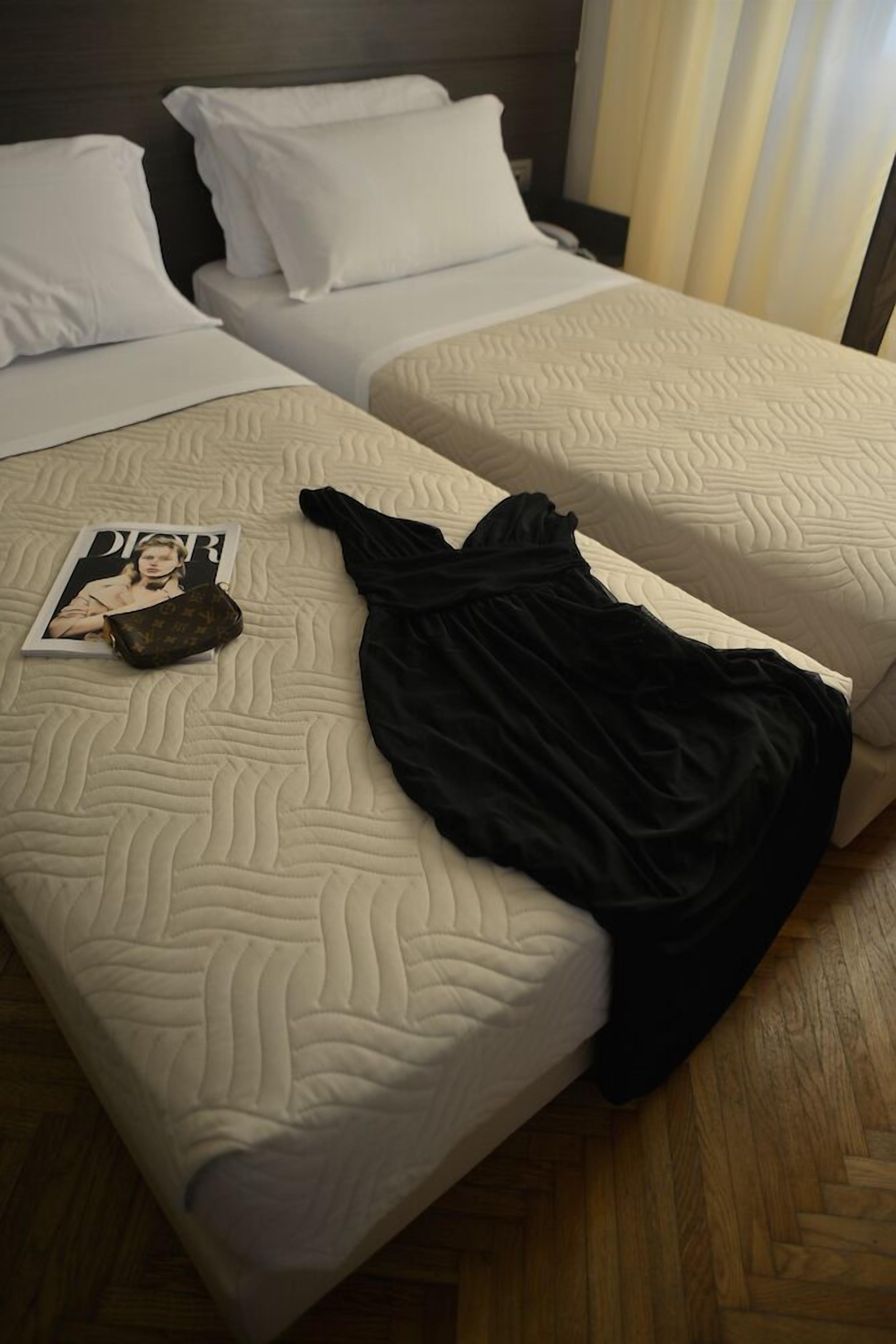 A hotel room with two beds, each with white bedding and a textured pattern. A black garment and a magazine are placed on the bed.