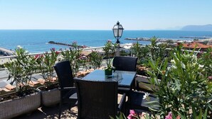 Un espacio al aire libre con sillas de mimbre y una mesa, con vistas a la costa, el mar y montañas distantes.