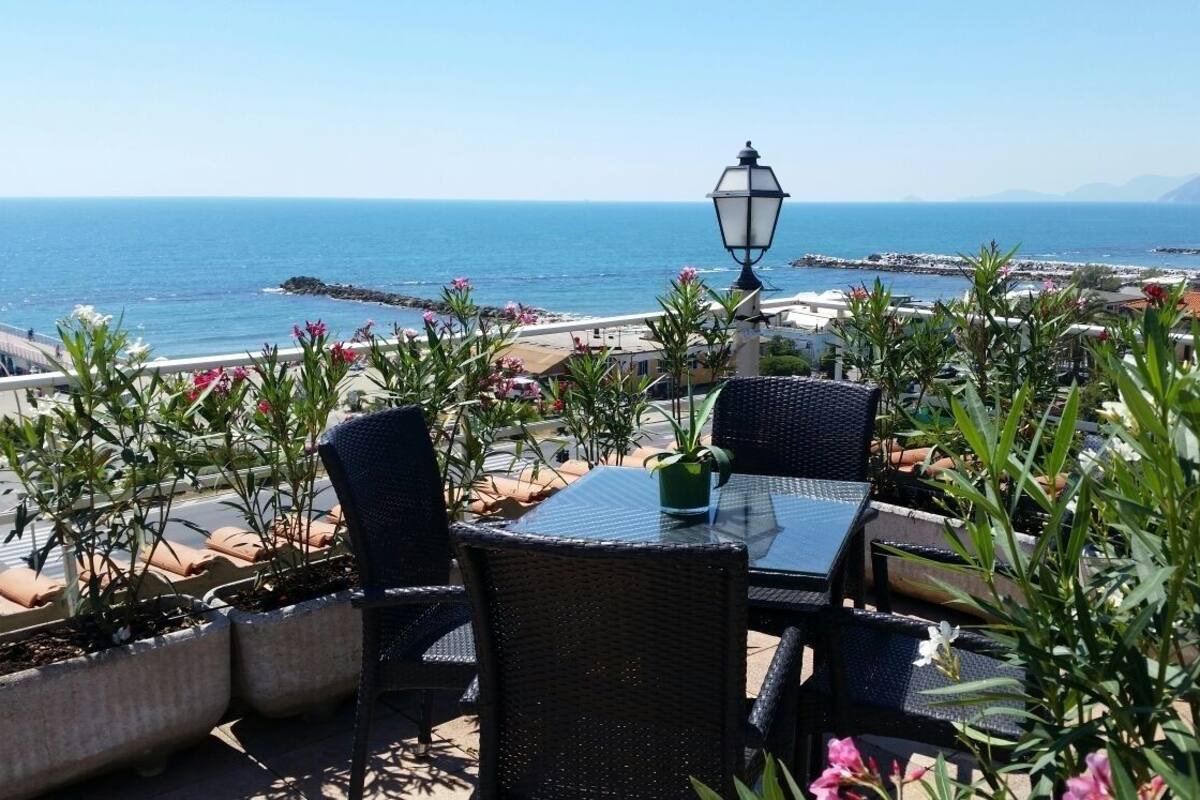 Un espace extérieur aménagé avec des chaises et une table en osier, offrant une vue imprenable sur la côte, la mer et les montagnes lointaines.