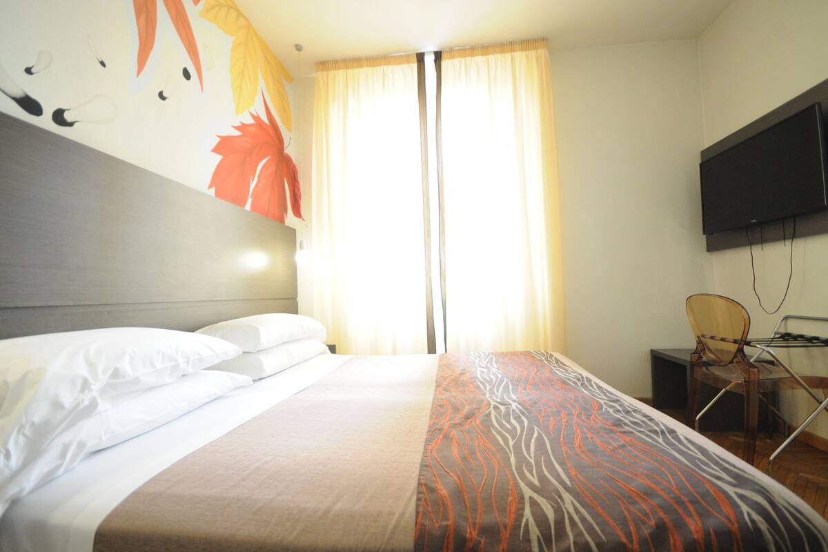 Une chambre d’hôtel comprenant un lit, un téléviseur à écran plat fixé au mur, un bureau avec une chaise et une fenêtre avec des rideaux.