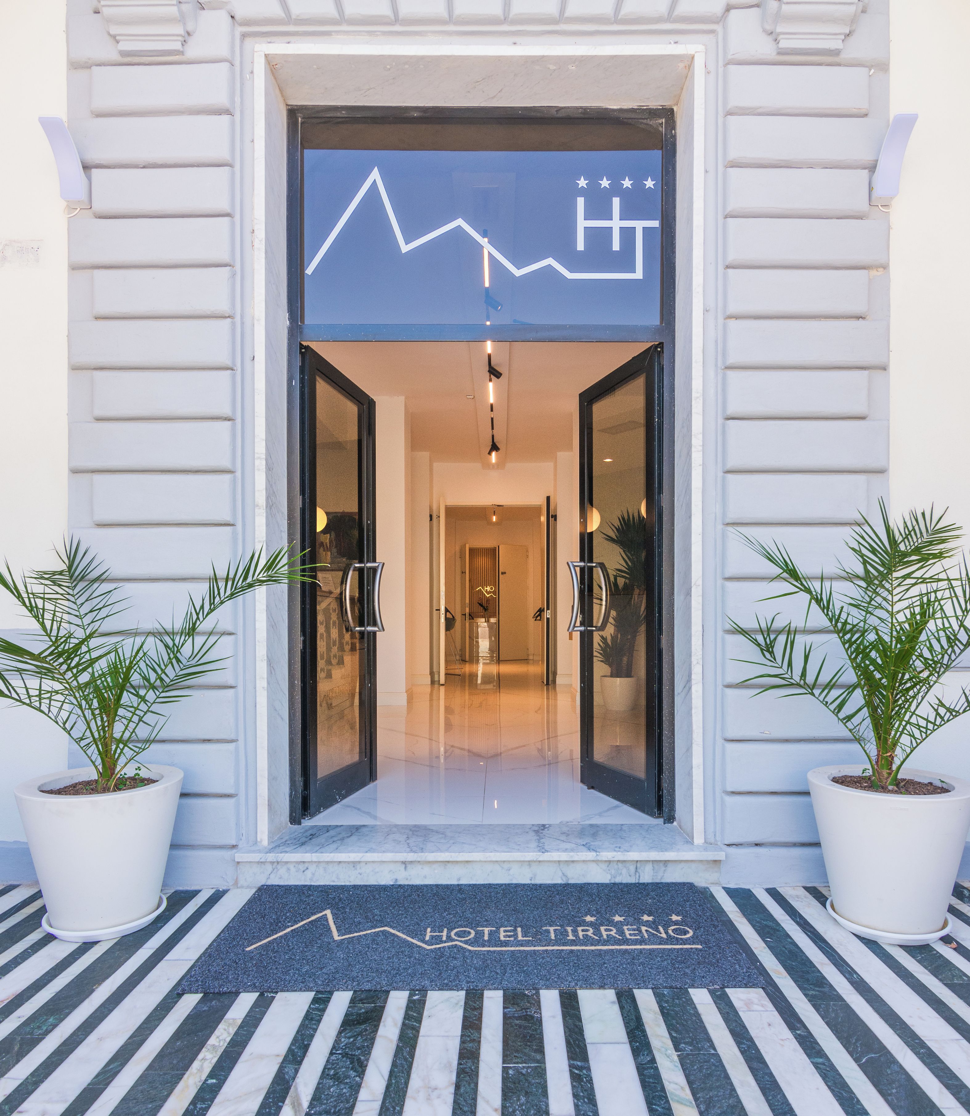 Entrada de hotel con diseño moderno, que incluye una puerta de cristal, un felpudo y plantas en macetas a ambos lados.