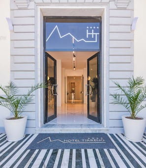 A hotel entrance with a modern design, featuring a glass door, a welcome mat, and potted plants on either side. - Hotel Tirreno (Massa)