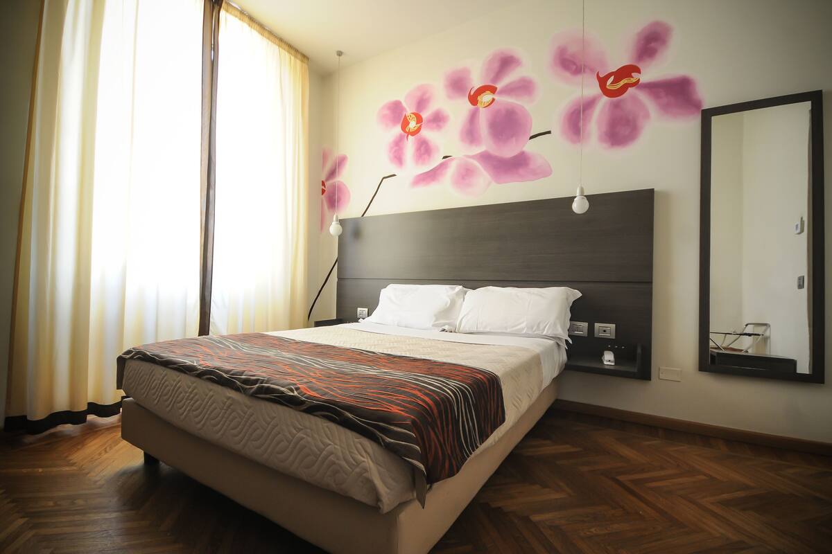 Une chambre d’hôtel avec un lit, une table de chevet, un miroir et un mur orné d’une fresque florale.