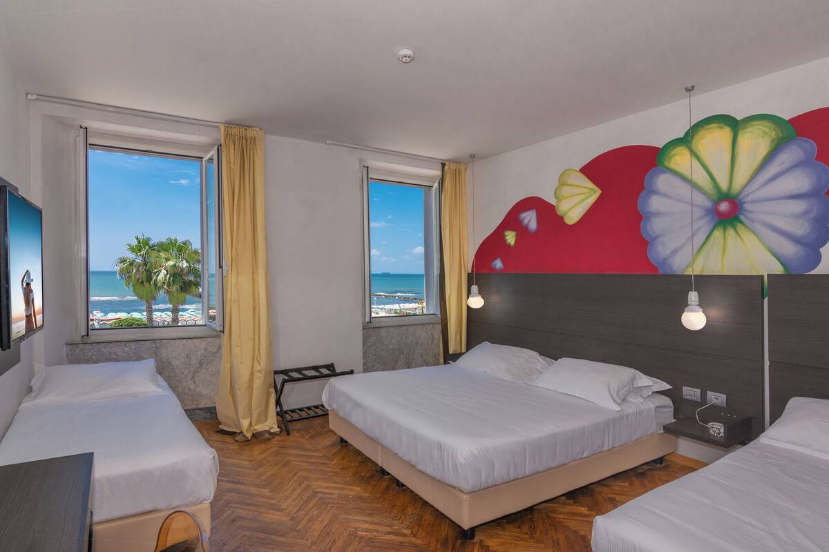 Une chambre d’hôtel avec deux lits, une grande fenêtre donnant sur la plage et une fresque colorée sur le mur.