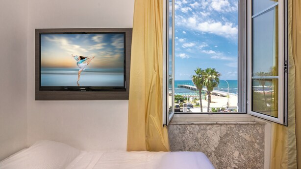 Una habitación de hotel con cama, televisor montado en la pared y ventana con vista a la playa y palmeras.