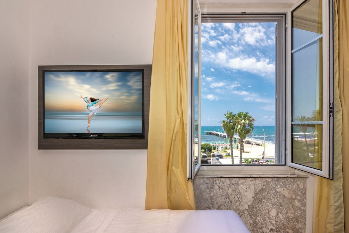 A hotel room with a bed, a television mounted on the wall, and a window with a view of the beach and palm trees.