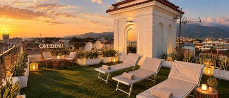 Una terraza en la azotea con sillones de descanso, plantas en macetas y una pequeña estructura blanca con techo rojo.