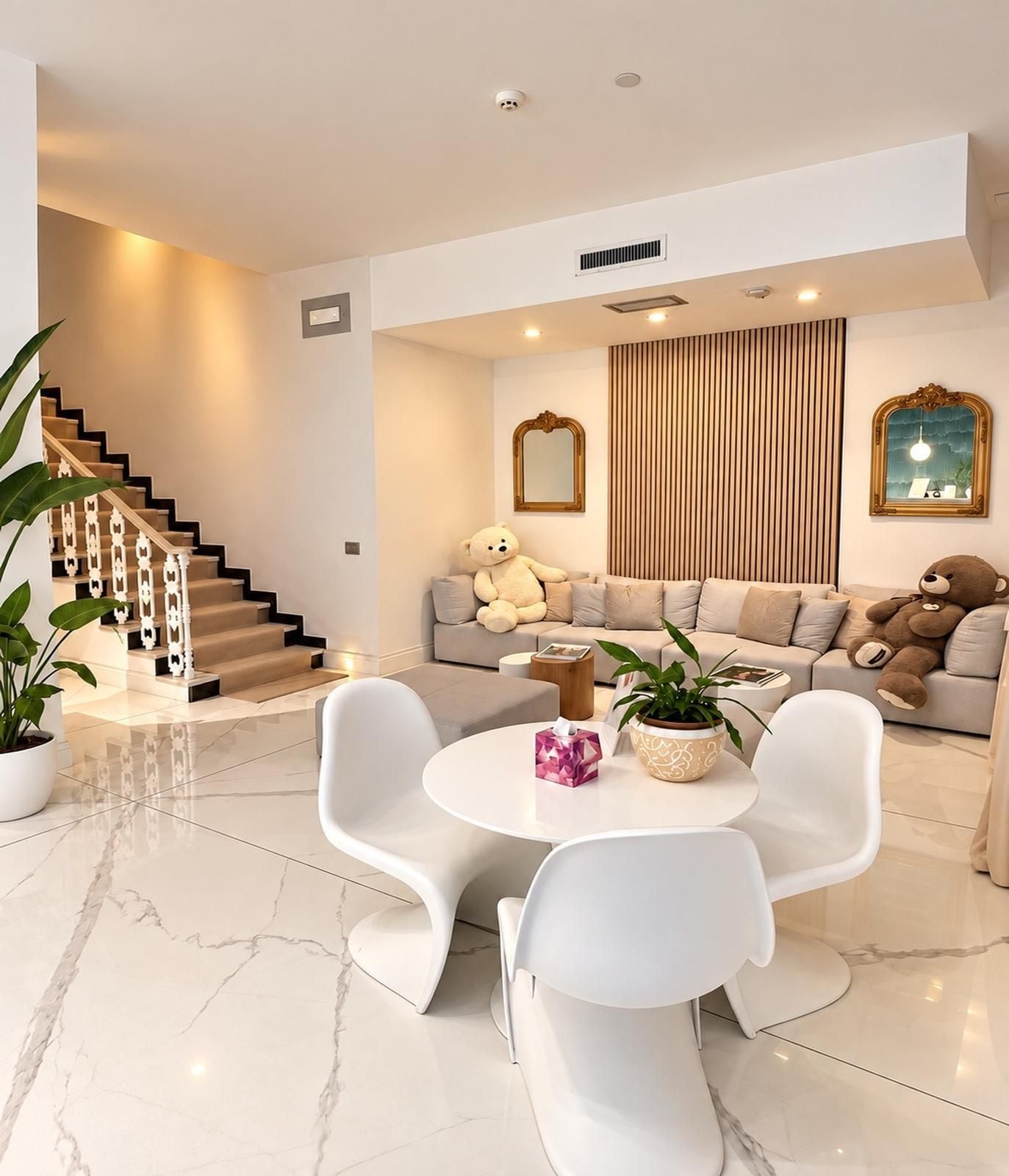 A modern living room with a staircase, a sofa set, two white chairs, a round table, and decorative elements like a mirror and plants.
